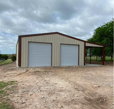A beige metal building with two garage doors and a covered area to the right, situated on a dirt lot.