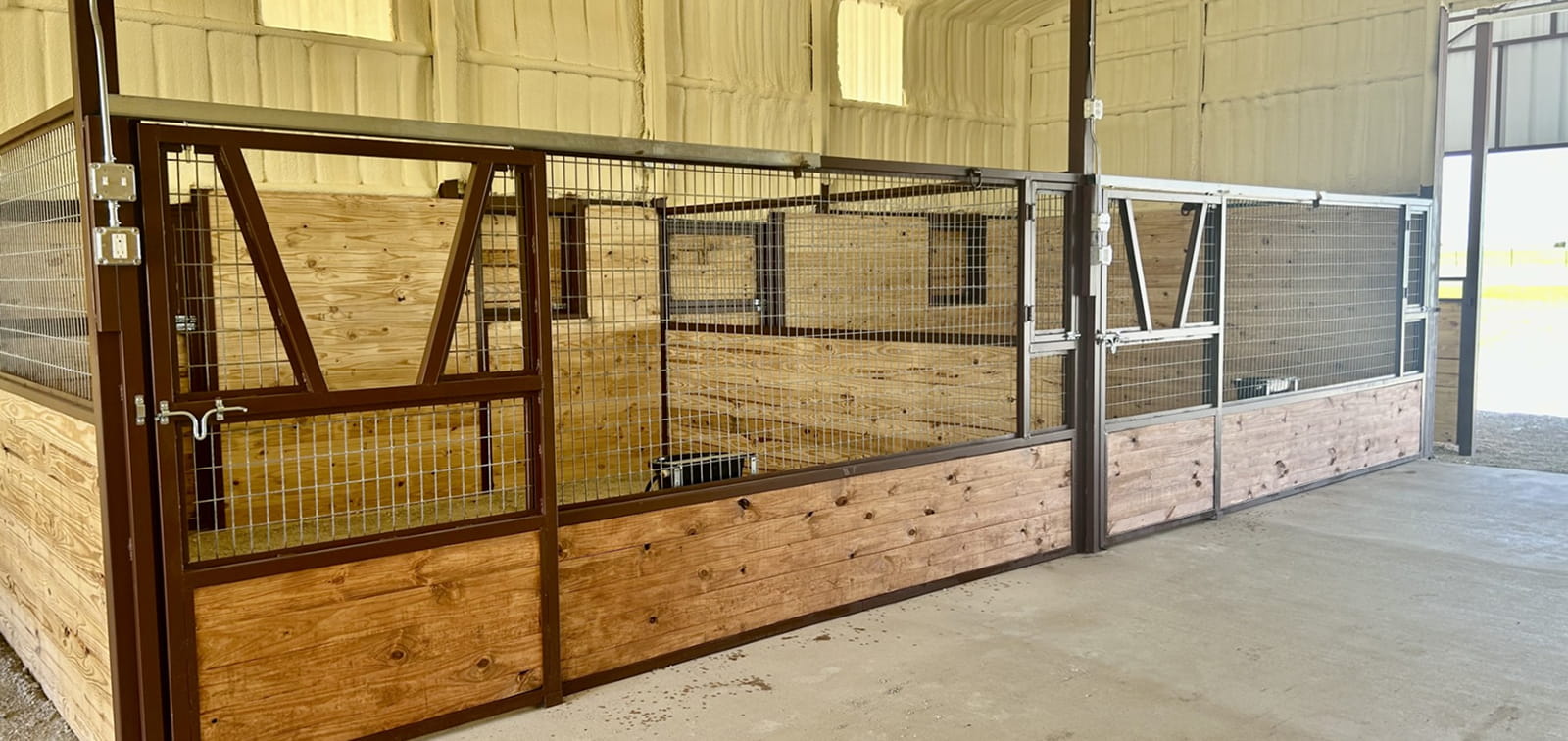 Three horse stalls with metal gates and wire mesh walls inside a barn.